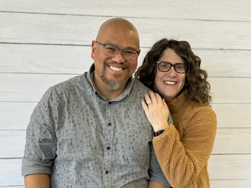 Smiling man with glasses and a goatee, wearing a patterned gray shirt, standing next to a smiling woman with curly hair, glasses, wearing a brown fleece jacket, against a white wooden wall.