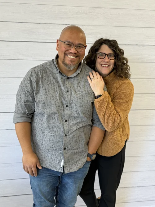 Smiling man with glasses and a goatee, wearing a patterned gray shirt, standing next to a smiling woman with curly hair, glasses, wearing a brown fleece jacket, against a white wooden wall.